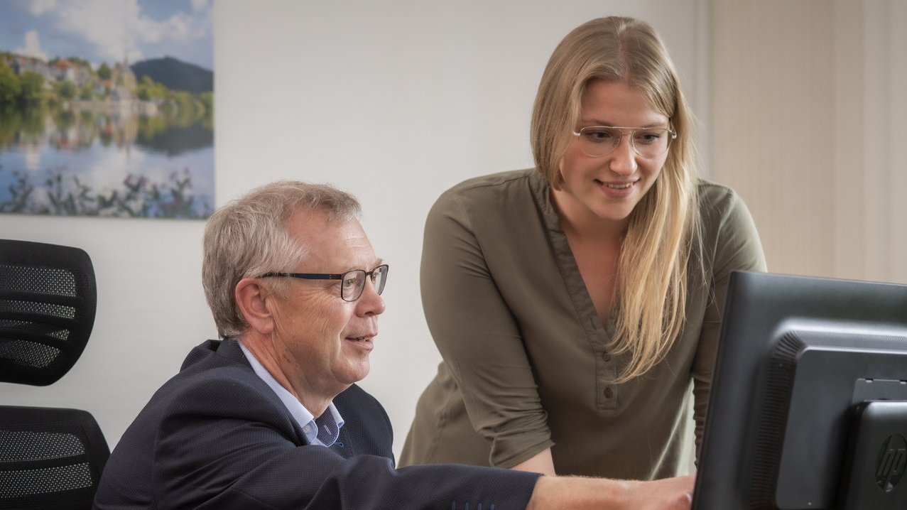 Ein Mann sitzt an einem Schreibtisch und eine Frau steht daneben.